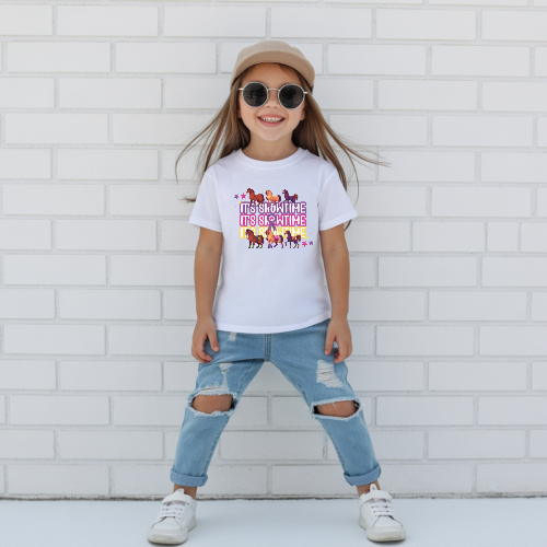 Child wearing a white t-shirt with colorful horse graphics and text, standing against a white brick wall.